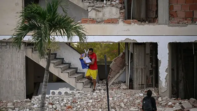 A man in yellow shorts and red shirt carries personal belongings after climbing down the steps of a partially collapsed building surrounded by debris