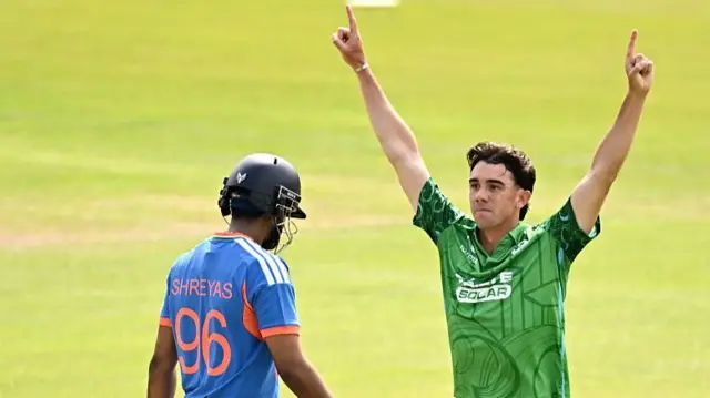 Matt Hollard (right), wearing a green Ireland shirt, raises both hands in the air and points his index fingers to the sky after taking the wicket of Shreyas Iyer (left), who is walking back past him wearing a blue India shirt and dark blue helmet