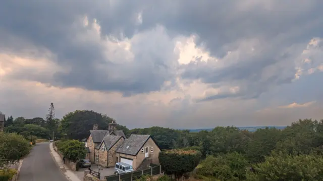 Dark clouds in the sky hang above a house which is surrounded by trees with an empty road in front of it
