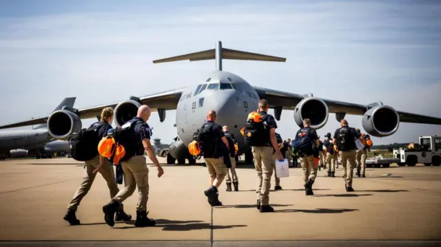 Emergency workers in brown trousers and blue shirts with orange helmets slung over their backs walk across the tarmac onto a plane.