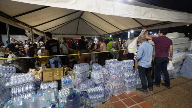 Two men stand outside a tent filled with crates of bottled water. Several people stand below the structure.