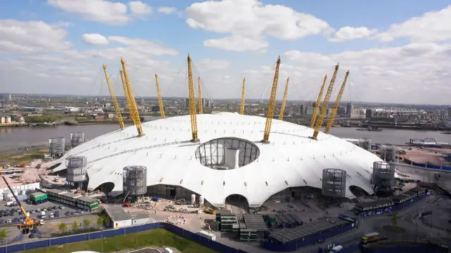 An aerial view of the O2 which is white and has long yellow metal structures coming up out of it