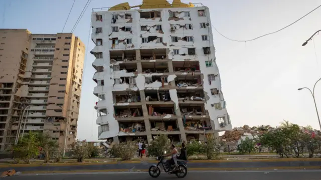Two men on a motorbike ride past a destroyed building on the verge of collapse