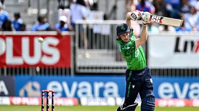 Lorcan Tucker, wearing a green Ireland shirt, blue helmet, and blue trousers and pads, plays a shot with the bat raised above his head