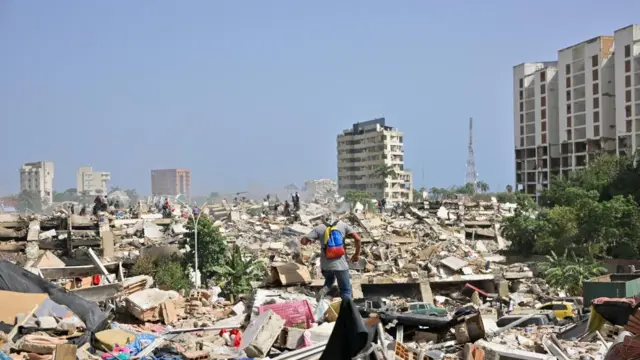 A man with a Venezuelan flag-themed backpack walks over the rubble of collapsed buildings in La Guaira several damaged towers stand in the backdrop