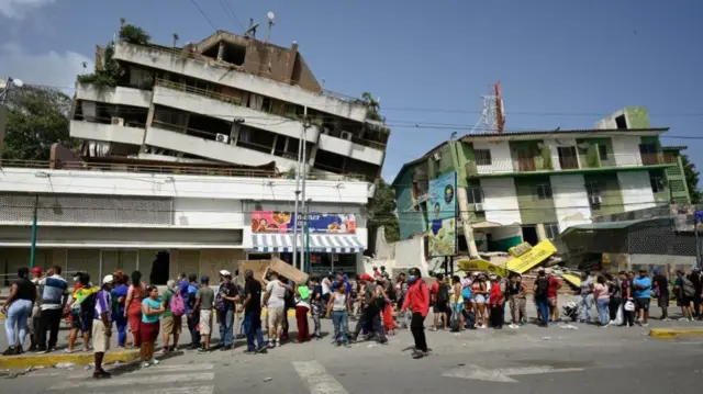 A line of people outside a shop waiting to receive aid, partially collapsed buildings are visible in the background.