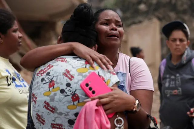 A woman waits, hugging another woman, as rubble is searched for survivors in La Guaira