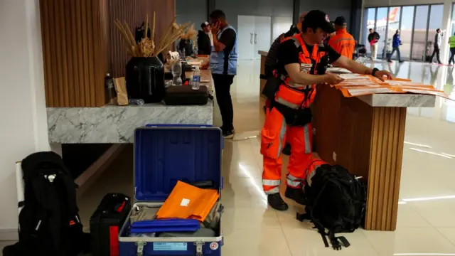 A man in high vis clothing looks at pieces of paper on a table; there is a bag near his foot