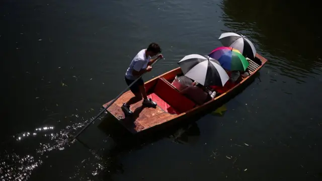 Three people shield themselves from the sun using umbrellas while punting in Cambridge. A man steers the end of the boat using an oar