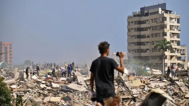 A boy stands filming a landscape of rubble on his phone