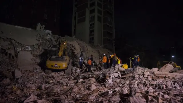 Several rescue workers stand on top of a pile of rubble next to a yellow digger
