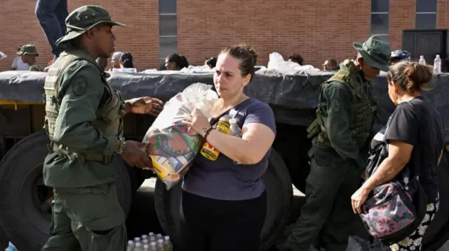 A woman holds a bag of groceries, surrounded by military personnel