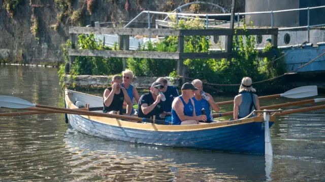 A group of six rowers pause for a water break. A seventh person sits at the end of the boat as the group drink bottles of water