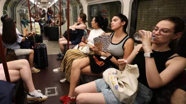 Commuters drink water and use electric and hand fans to try and stay cool on the Tube in London