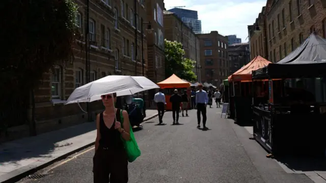 Woman walking underneath an umbrella and with sunglasses on through street lined with market food stalls