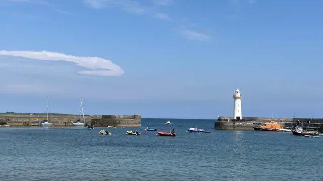A view over a harbous showing boats moored in the water and a lighthouse. The sky is blue and mostly clear with some clouds