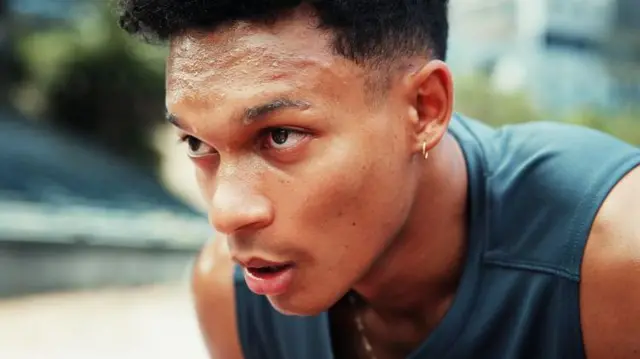A young man, wearing a sports vest top, is out for a run. He is hunched over, hands on knees, sweating