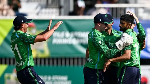 Jai Moondra (right) is congratulated by three teammates, all wearing green Ireland shirts and dark blue trousers, with Matt Hollard running in from the left with both hands raised