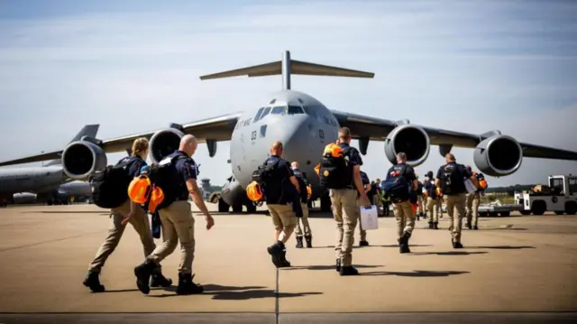 About a dozen rescue workers dressed in tan trousers and blue shirts, with orange helmets, walk across the tarmac onto a plane