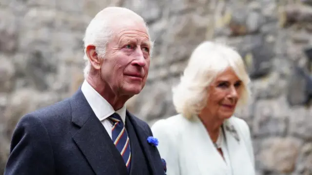King Charles III wears a navy suit and striped tie, next to him Camilla is wearing a white jacket, they stand in front of a stony wall outside