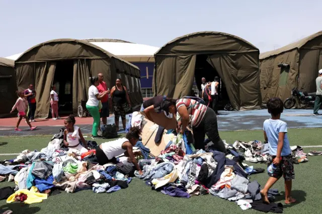 Rows of brown tents, in front on the grass is a pile of clothes and people