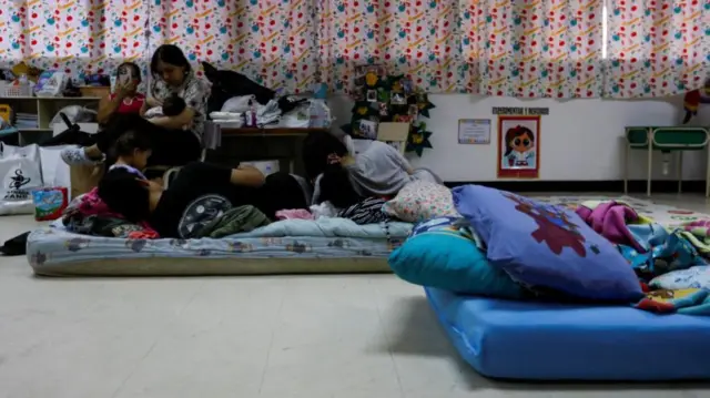 Two mattresses on the floor of a classroom, some children sit together on the first mattress