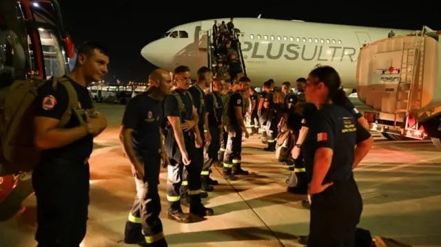 Rows of rescue workers stand on the tarmac by a plane at night time
