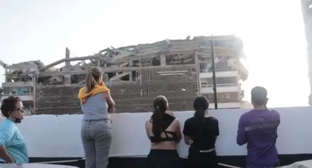 A row of people stand and survey a collapsed building