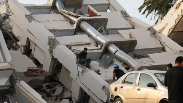 A collapsed building lies on its side, with a damaged car in front of it