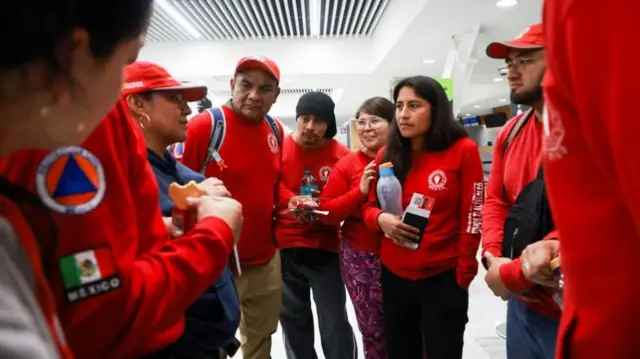 A group of people wearing red sweatshirts and caps gather in a huddle in a white-walled room
