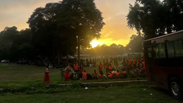 A large group of emergency workers in high-visibility jackets stand on some grass at sunset