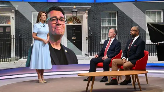 Victoria Derbyshire wears a blue dress and stands in a TV studio set, sat down next to her are Steve Reed and James Cleverly