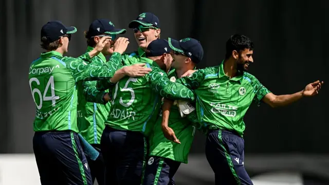 Ireland's players, wearing green shirts, dark blue trousers, and dark blue caps, embrace in a group celebration