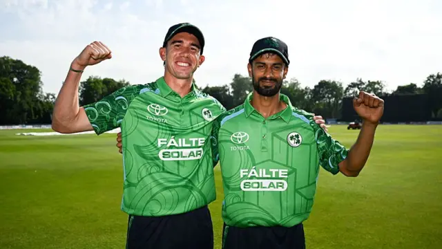 Matt Hollard (left) and Jai Moondra (right), wearing green Ireland shirts, dark blue trousers and dark blue caps, stand arm in arm with their fists raised in celebration