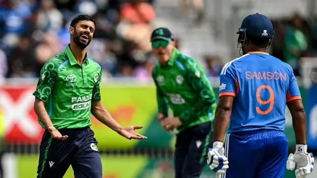 Jai Moondra, wearing a green Ireland shirt and dark blue trousers, smiles and holds his arms outstretched in front of him after dismissing Sanju Samson (right), wearing blue shirt and trousers and holding a bat in his left hand