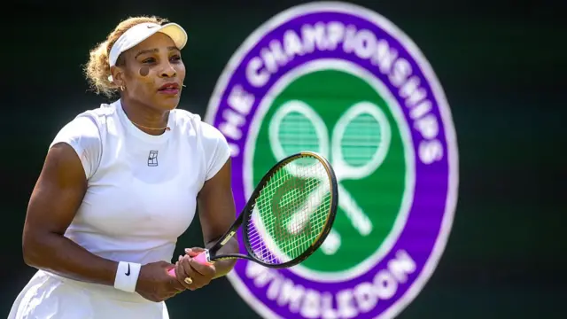 Serena Williams (left), wearing a white dress and cap, holds her racquet in two hands with the Wimbledon logo (purple circle with The Championships Wimbledon in white text around a green circle with two racquets crossed over each other in white)