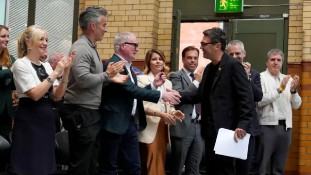 A row of people stand up to greet Andy Burnham.One of them, a man in a blazer and white shirt, is shaking Andy Burnham's hand while the others around him are clapping.