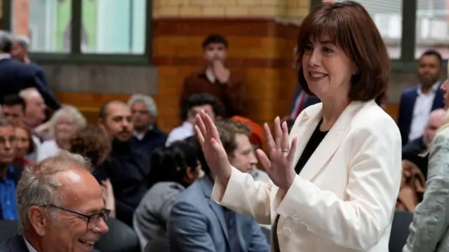 Lucy Powell stands up and raises her hands to people in a crowd sat in front of her. She has long brown hair and is wearing a white blazer over a black top.