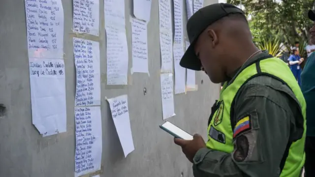 A man in police uniform holds his phone and looks at long lists with names stuck to a wall