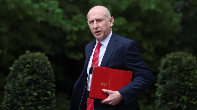 John Healey walks in a navy suit with a red tie in. He is holding a red folder.
