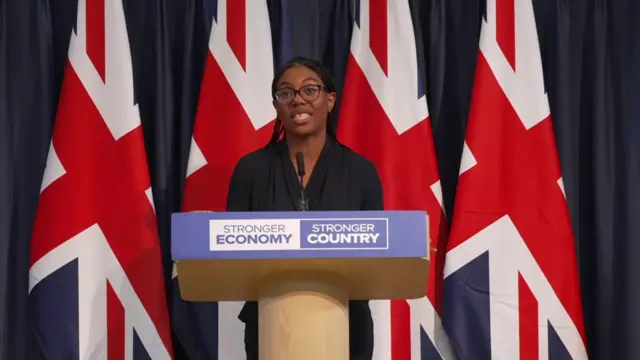 Kemi Badenoch speaking behind a lectern which reads stronger economy, stronger country. Four Union Jack flags hang behind her