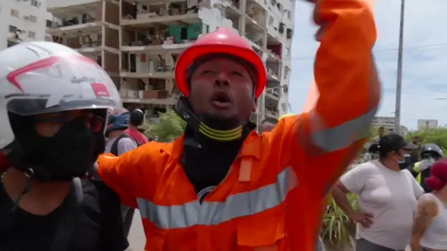Wilker Molaya in an orange hi-vi jacket clamours as he holds on to the back of a woman in an helmet, a severely damaged building behind him