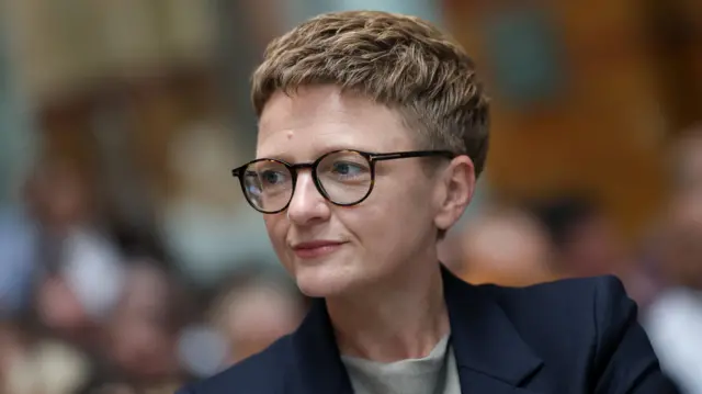 Bev Craig, who has short blonde hair, is wearing tortoise shell coloured glasses, a navy blazer and grey top underneath. She is standing in front of a seated crowd who appear blurry in the background of the photo