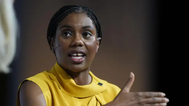 Kemi Badenoch, a woman, wearing yellow, with her right hand pointing towards the left.