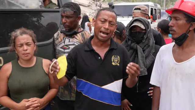 A man in a black polo shirt speaks to Reuters in front of a group of people that have been searching through the rubble to find survivors of earthquakes