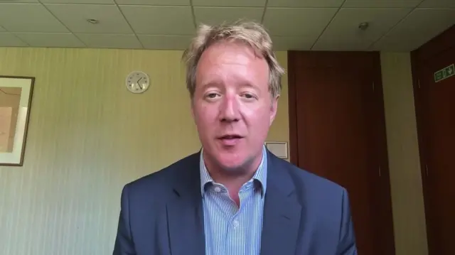 Paul Bristow wears a navy suit while sitting in what appears to be a hotel room. A clock is on the wall behind him.