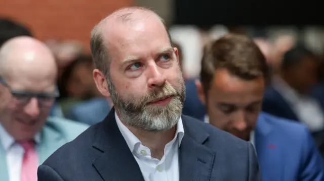 Jonathan Reynolds is straight faced and looks upwards while sat on a chair in the front row of a seated crowd. He is bald with a grey and black short beard. He is wearing a navy blazer and white shirt without a tie.