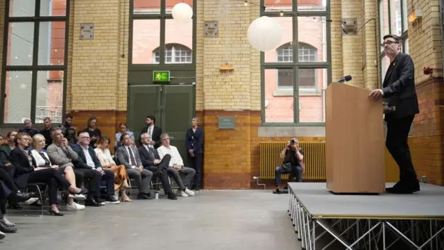 Andy Burnham stands on a stage and speaks to politicians, activists and journalists sat on chairs in front of him