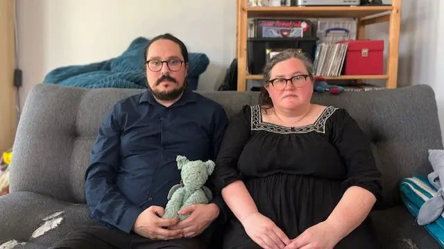 A man and woman sat on a grey sofa, the man holds a child's cuddly toy