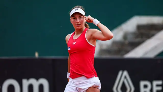 Teodora Kostovic, wearing a red vest and white visor, points her left index finger to her temple
